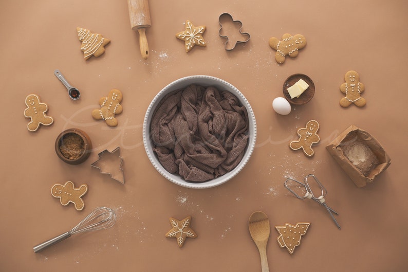 May include: A brown background with a variety of gingerbread cookies, cookie cutters, and baking supplies. There is a brown cloth in a white bowl in the center of the image.