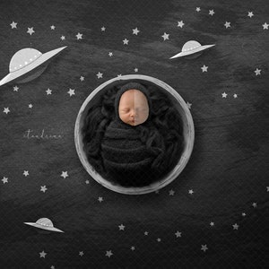 May include: A newborn baby is swaddled in a dark gray fabric and sleeping in a white bowl. The baby is surrounded by white stars and flying saucers on a black background.
