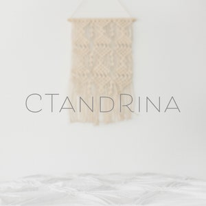 May include: A white, fluffy bedspread with a macrame wall hanging above it. The wall hanging is made of natural fibers and has a geometric pattern. The text "CTANDRINA" is printed in gray on the wall below the hanging.