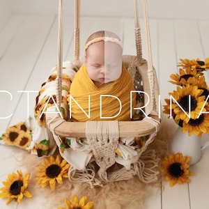 May include: A macrame swing seat with a wooden frame containing a swaddled infant wearing a headband. The scene includes a sunflower-patterned blanket, a white pitcher filled with sunflowers, and loose sunflowers. The text CTANDRINA is visible.