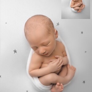May include: A newborn baby is swaddled in a white blanket and is sleeping on a white background with grey stars.