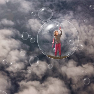 May include: A young girl in a pink skirt and grey sweater blows bubbles in a large, clear bubble. The girl is standing in the center of the bubble, which is surrounded by smaller bubbles.