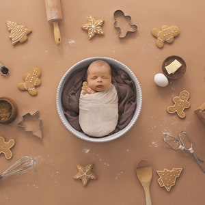 May include: A newborn baby is swaddled in a white blanket and lying in a white bowl. The baby is surrounded by gingerbread cookies, cookie cutters, and baking supplies, including a rolling pin, a whisk, a wooden spoon, a bowl of brown sugar, a bag of flour, and an egg.