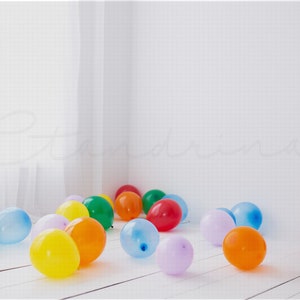 21 total Orange, purple, blue, red, and green balloons that represent the rainbow, laying on a white wooden plank floor inside of a clean white studio with the light from the window illuminating into the room.