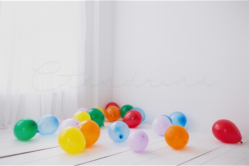 21 total Orange, purple, blue, red, and green balloons that represent the rainbow, laying on a white wooden plank floor inside of a clean white studio with the light from the window illuminating into the room.