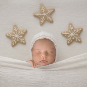 May include: A newborn baby wearing a white bonnet, resting peacefully on a white blanket. Three star-shaped cookies with white icing are arranged above the baby. The image has a soft, neutral color palette.