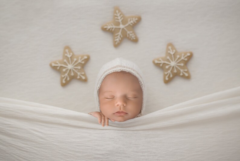 May include: A newborn baby wearing a white bonnet, resting peacefully on a white blanket. Three star-shaped cookies with white icing are arranged above the baby. The image has a soft, neutral color palette.