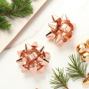 May include: Two rose gold starburst bows, with a metallic finish, are displayed on a white surface, alongside gold ornaments and green pine branches. These decorative bows are ideal for gift wrapping or holiday decor.