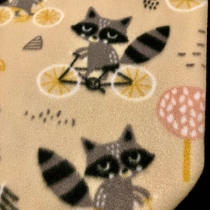 May include: A beige fabric featuring a repeating pattern of raccoons riding bicycles. The raccoons are gray and black with white accents, and the bicycles have yellow wheels. The background includes stylized trees and dots.