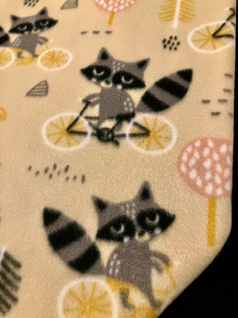 May include: A beige fabric featuring a repeating pattern of raccoons riding bicycles. The raccoons are gray and black with white accents, and the bicycles have yellow wheels. The background includes stylized trees and dots.