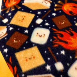 May include: A navy blue fabric with a repeating pattern of s'mores, campfires, and stars. The s'mores have smiling faces and are made of graham crackers, chocolate, and marshmallows. The campfires are orange and yellow.