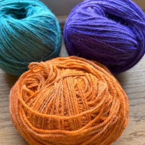 May include: Three balls of yarn in different colours: teal, purple, and orange. The yarn is wound into balls and ready for knitting or crocheting.