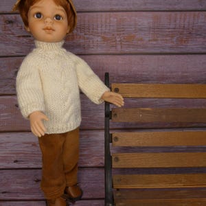 Peut inclure: Poupée vêtue d'un pull en tricot crème, d'un pantalon en velours côtelé marron et de chaussures en cuir marron. La poupée a les cheveux roux et porte un petit chapeau de paille. Elle se tient près d'un banc en bois.