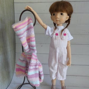 Peut inclure: Une poupée vêtue d'un haut blanc et d'une salopette à rayures roses, avec des bottines roses. Un pull tricoté à rayures multicolores est suspendu à un support en métal foncé. Le pull présente des rayures roses, jaunes et bleues.
