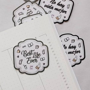 Könnte beinhalten: Ein weißer Planer mit einem gepunkteten Rastermuster und einem silbernen Aufkleber mit der Aufschrift "Best Life Ever" mit einem Kaffeetassen- und Briefumschlagdesign. Auf der Seite befinden sich weitere Aufkleber mit spanischem Text.