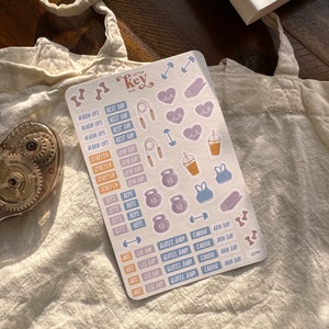 May include: A sheet of colorful fitness themed stickers with illustrations of dumbbells, jump ropes, hearts, and workout clothing. The stickers have text that includes "Warm-ups", "Rest Day", "Gym Day", "Sets", "Reps", "Abs", "Leg Day", "Glutes, Baby", "Cardio", and "Arm Day".