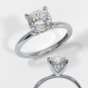 May include: A silver engagement ring featuring a cushion-cut diamond. The ring is set with a halo of smaller diamonds and has a simple band. The ring is engraved with the words "Art of Fine".