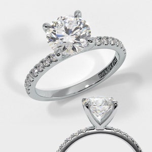 Könnte beinhalten: Ein silberner Verlobungsring mit einem großen, runden, klaren Diamanten als Mittelstein. Das Band ist mit kleineren Diamanten besetzt. Die Worte "ART OF FINE" sind in das Band eingraviert. Auf weißem Hintergrund.