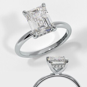 May include: A silver engagement ring featuring an emerald cut diamond with a halo of smaller diamonds. The ring is engraved with "ART OF FINE".