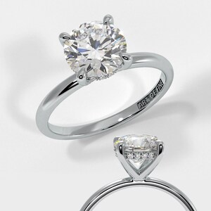 May include: A silver engagement ring featuring a round, clear diamond centre stone with a halo of smaller diamonds. The band is plain and the ring is engraved with "ART OF FINE".