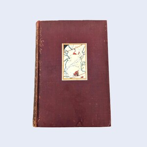 May include: A burgundy hardback book with a gold-trimmed illustration of a map on the front cover. The illustration features a small red figure on a white background.