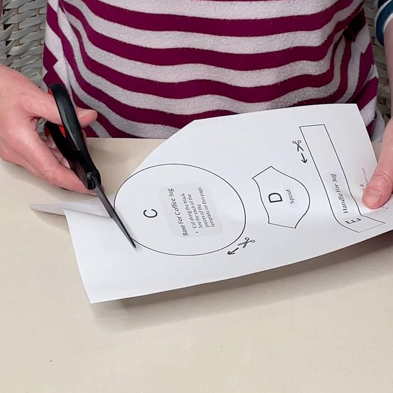 May include: Scissors cutting a paper template for a coffee jug. The template includes labeled sections for the base, spout, and handle. The paper is white with black printed lines and text. The person is wearing a striped shirt.