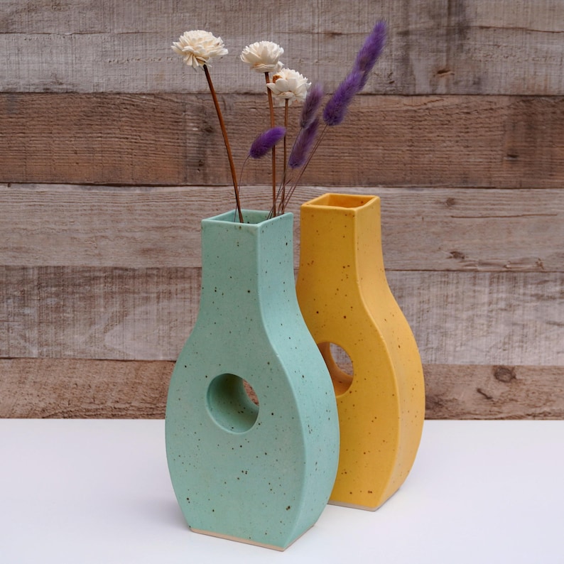 Op de afbeelding: Twee keramische vazen, &eacute;&eacute;n in lichtgroen en de andere in geel. Beide vazen hebben een unieke, asymmetrische vorm met een ronde opening aan de zijkant. De vazen zijn bezaaid met kleine witte stippen.