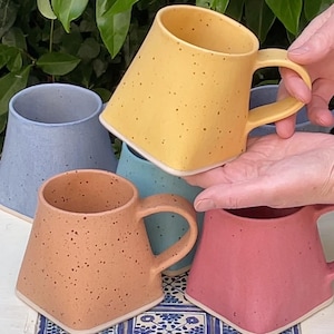 Pode incluir: Quatro canecas de cer&acirc;mica com esmalte salpicado em diferentes cores: amarelo, azul, azul claro e rosa. As canecas s&atilde;o de forma quadrada com uma al&ccedil;a arredondada.