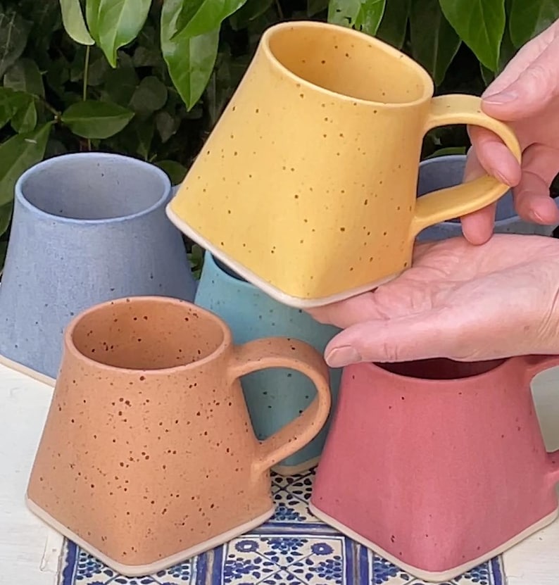Pode incluir: Quatro canecas de cer&acirc;mica com esmalte salpicado em diferentes cores: amarelo, azul, azul claro e rosa. As canecas s&atilde;o de forma quadrada com uma al&ccedil;a arredondada.