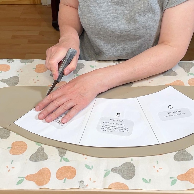 May include: A person using a craft knife to cut around a paper template labeled "Teapot Side" on a tan material. The cutting is taking place on a patterned fabric with pears.