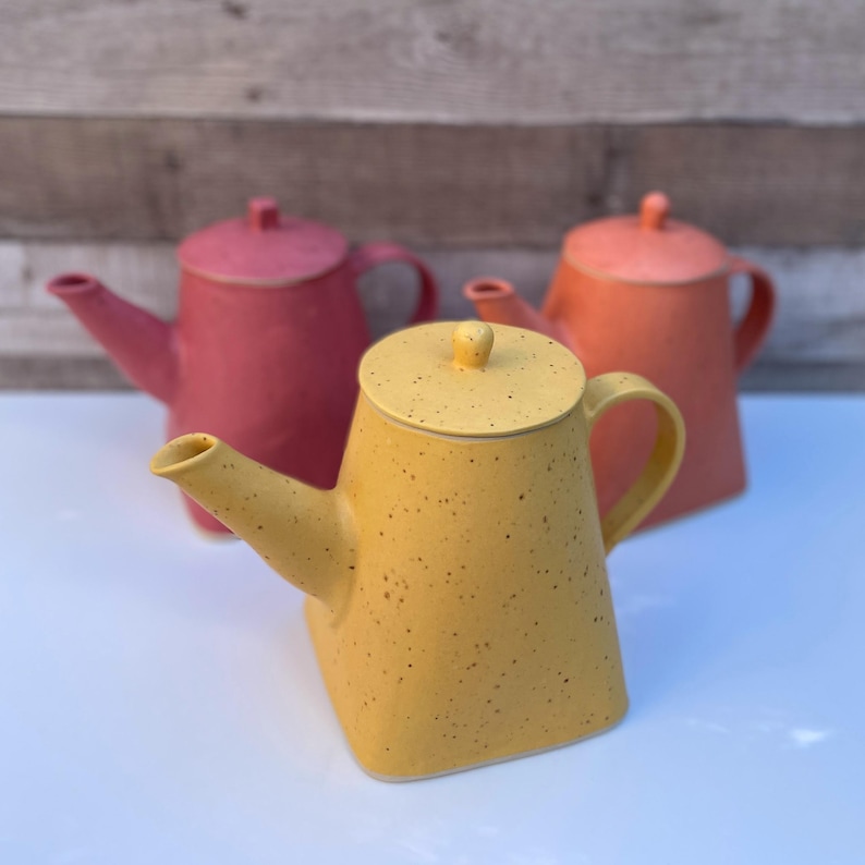 May include: Three ceramic teapots in shades of yellow, coral, and pink. The yellow teapot in the foreground has a speckled finish. Each teapot has a lid and a curved handle. The teapots are arranged on a white surface.