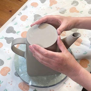 May include: A light gray ceramic teapot with a matching lid. The teapot has a handle and a spout. The teapot is being held by two hands. The teapot is sitting on a clear, round surface.