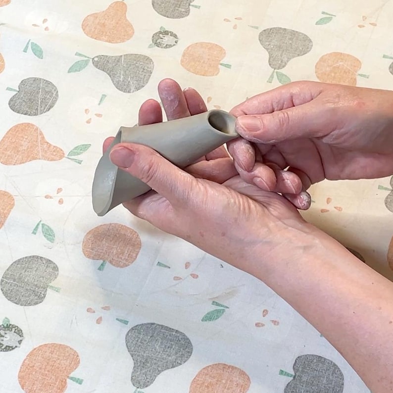 May include: A gray ceramic funnel-shaped object with a wide base and a narrow spout is held between two hands. The object is unglazed and appears to be handmade. The background is a patterned fabric with fruit designs.