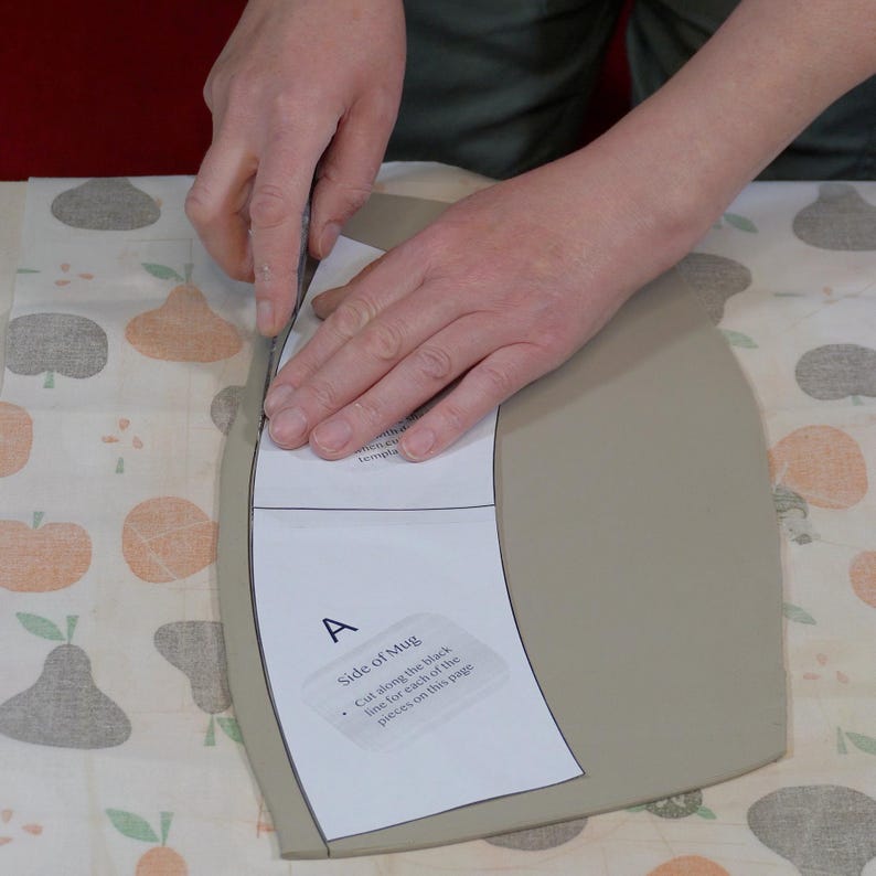 May include: A person cuts a piece of clay with a knife. The clay is shaped like a mug and has a paper template attached to it. The template has instructions for cutting the clay along the black line for each of the pieces on the page.
