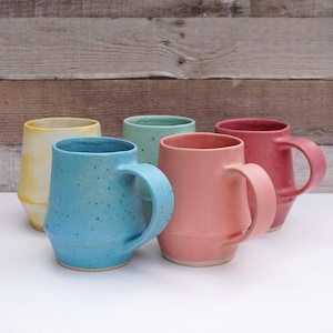 May include: Five ceramic mugs in different colors, including yellow, light blue, green, pink, and red. The mugs have a unique, slightly rounded shape with a handle on the side. The mugs are arranged in a row on a white surface.