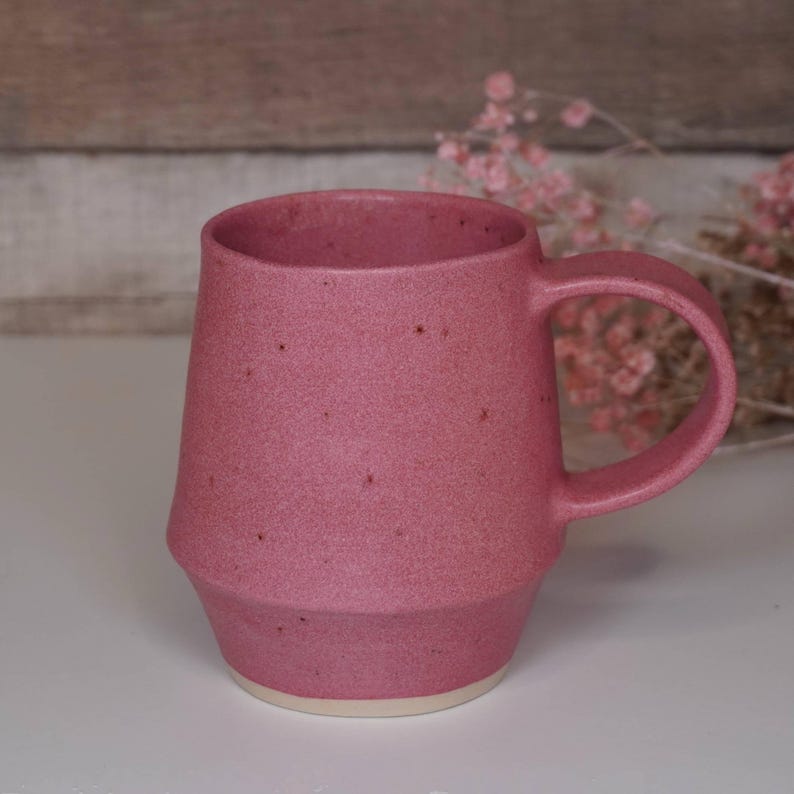 May include: A speckled pink ceramic mug with a rounded body and a handle. The mug has a light brown rim.
