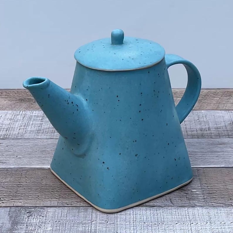 May include: A turquoise ceramic teapot with a square base and a matching lid. The teapot has a curved handle and a spout. The surface has a speckled texture. The teapot is a light blue color.