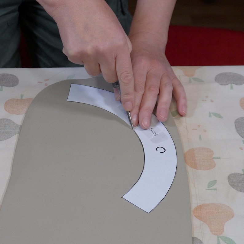 May include: A person uses a craft knife to cut a paper pattern into a piece of gray clay. The pattern is labeled "C" and "trim us".