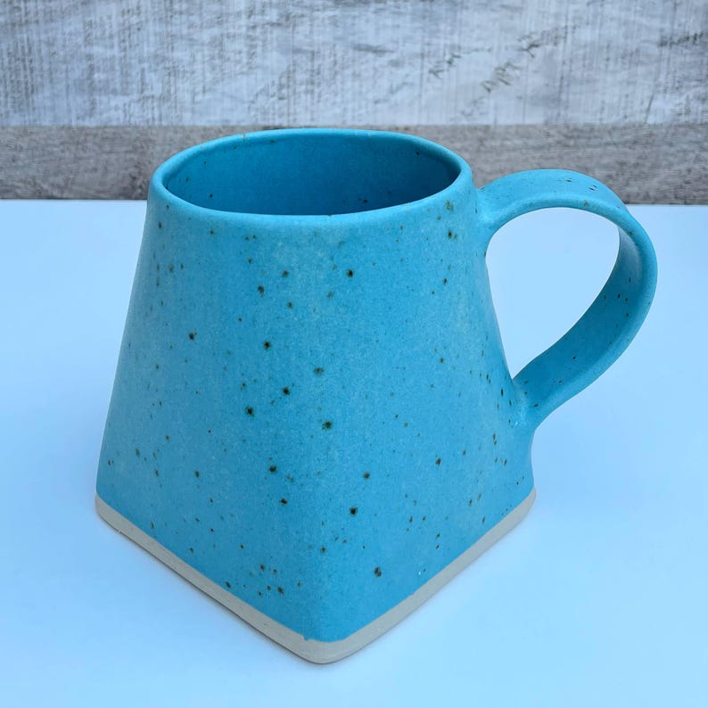 Pode incluir: Uma caneca de cer&acirc;mica azul turquesa com um esmalte salpicado e uma borda branca. A caneca tem uma base quadrada e uma al&ccedil;a arredondada.