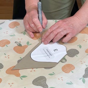 May include: A person is using a craft knife to cut a tan-colored piece of material. A paper template labeled "Teapot Spout" is placed on the material. The background features a patterned fabric with illustrations of pears.