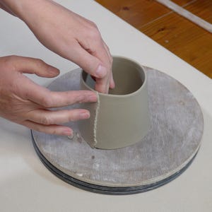 May include: A person's hands are shown shaping a light brown clay pot on a pottery wheel. The pot is being formed into a cylindrical shape with a slightly flared top.