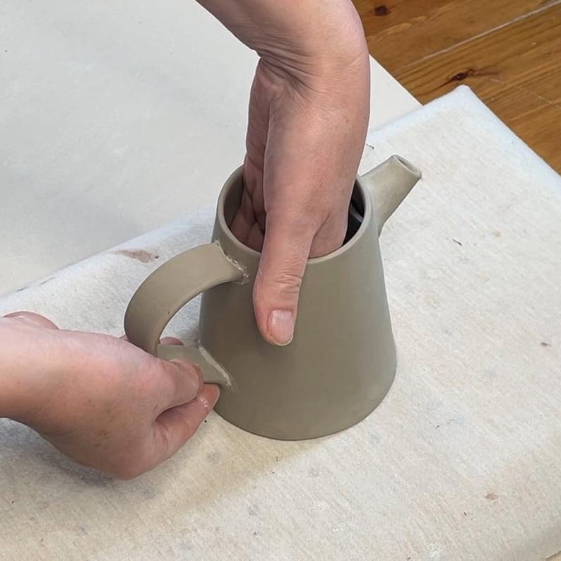 May include: A beige ceramic teapot with a curved handle and a spout. The teapot is being held by two hands. The teapot has a smooth, matte finish and a simple, modern design.