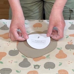 May include: A person's hands are holding a paper template labeled "Teapot Lid" over a piece of clay. The template is white with black text and a black outline. The clay is a light brown color, and the background is a patterned fabric.