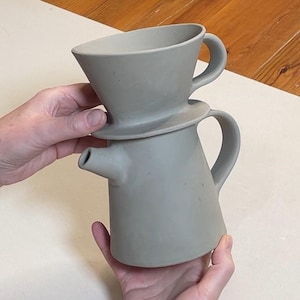 May include: A two-piece, light gray ceramic coffee maker. The top piece is a cone-shaped pour-over with a handle. The bottom piece is a pitcher with a spout and handle. The coffee maker is held by two hands.