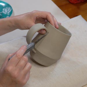 May include: A person is using a tool to trim the edge of a ceramic mug with a handle. The mug is light brown and has a rounded shape.