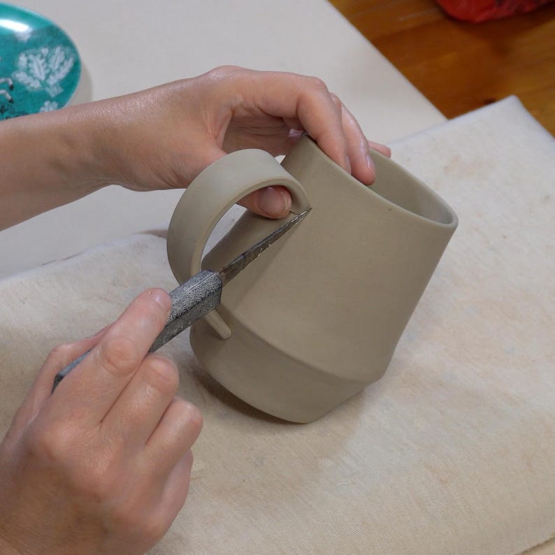 May include: A person is using a tool to trim the edge of a ceramic mug with a handle. The mug is light brown and has a rounded shape.