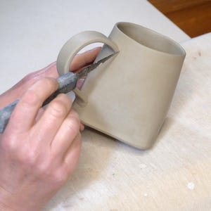 Pode incluir: Uma m&atilde;o usa uma faca pequena para aparar a al&ccedil;a de uma caneca de cer&acirc;mica bege.