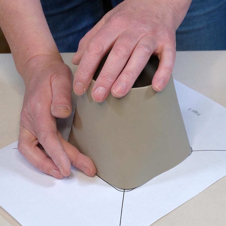 Pode incluir: Uma pessoa est&aacute; usando um modelo para moldar um peda&ccedil;o de argila. A argila &eacute; de cor marrom claro e est&aacute; sendo moldada em um cone. O modelo &eacute; uma folha de papel branca com linhas pretas desenhadas nela.