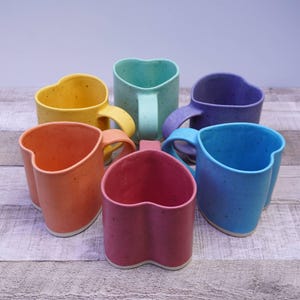 May include: Six heart-shaped ceramic mugs in various colors: yellow, orange, teal, purple, red, and blue. Each mug has a matching handle and a speckled texture. The mugs are arranged on a light-colored wooden surface.