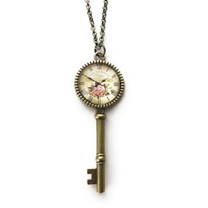 May include: A gold key necklace with a clock face pendant featuring Roman numerals and a floral design. The clock face says "Le Temps est d'Or" which translates to "Time is Gold".